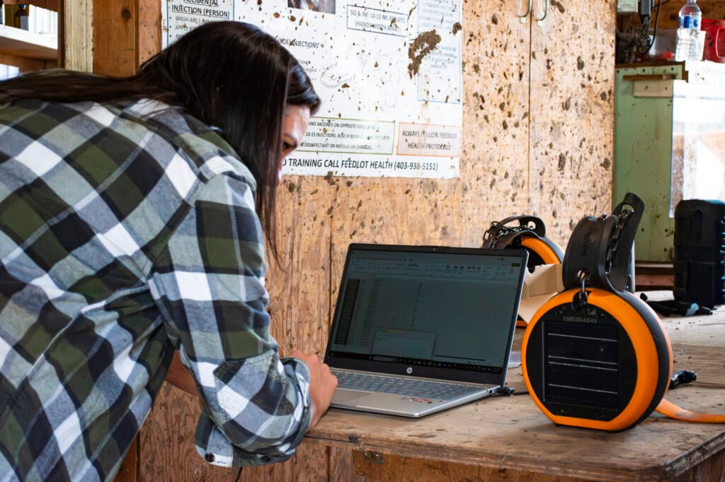 A person in a plaid shirt leaning over a wooden workbench to use a laptop beside an eShepherd GPS virtual fencing neckband, with livestock health posters on the wall behind them.